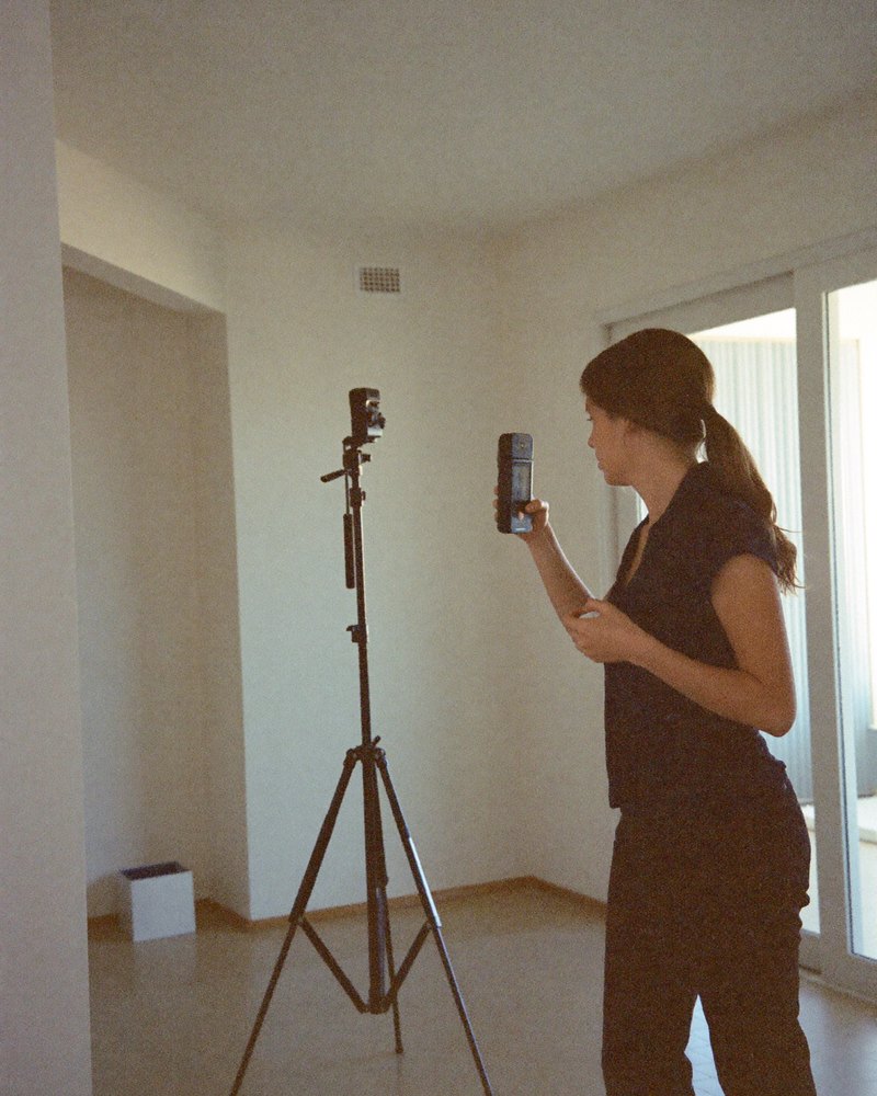 An American real estate agent in her 30s photographs an empty bright living room using a phone mounted on a small tripod.