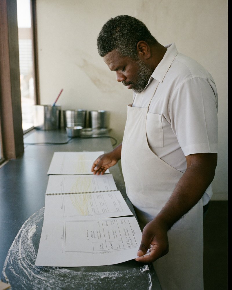 A Black American bakery owner in his 50s reviews printed website mockups spread across his counter.