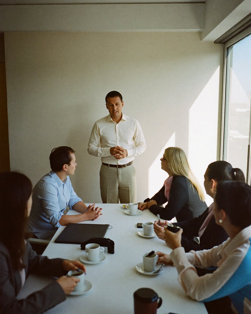 A small early-morning business breakfast meeting. A Latino man in his 30s stands to speak while others at the table listen, coffee cups in front of them.
